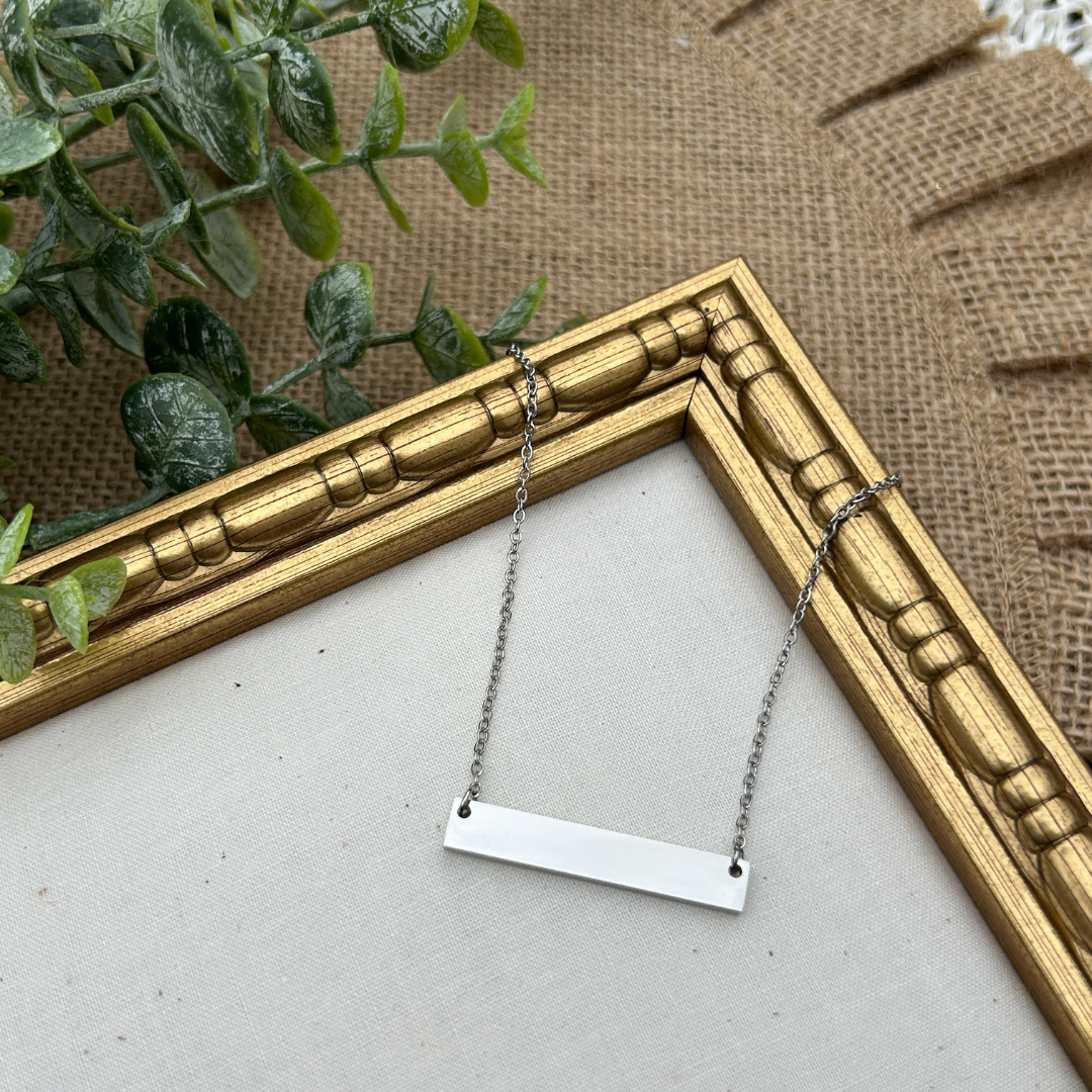 Silver bar necklace on a white surface with a gold frame and green plant in the background