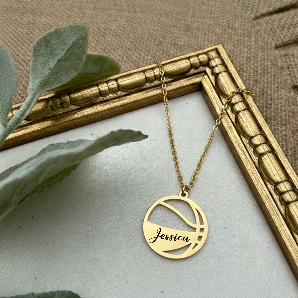 Gold necklace with 'Jessica' on the basketball pendant on a textured surface with a gold frame and green leaves.
