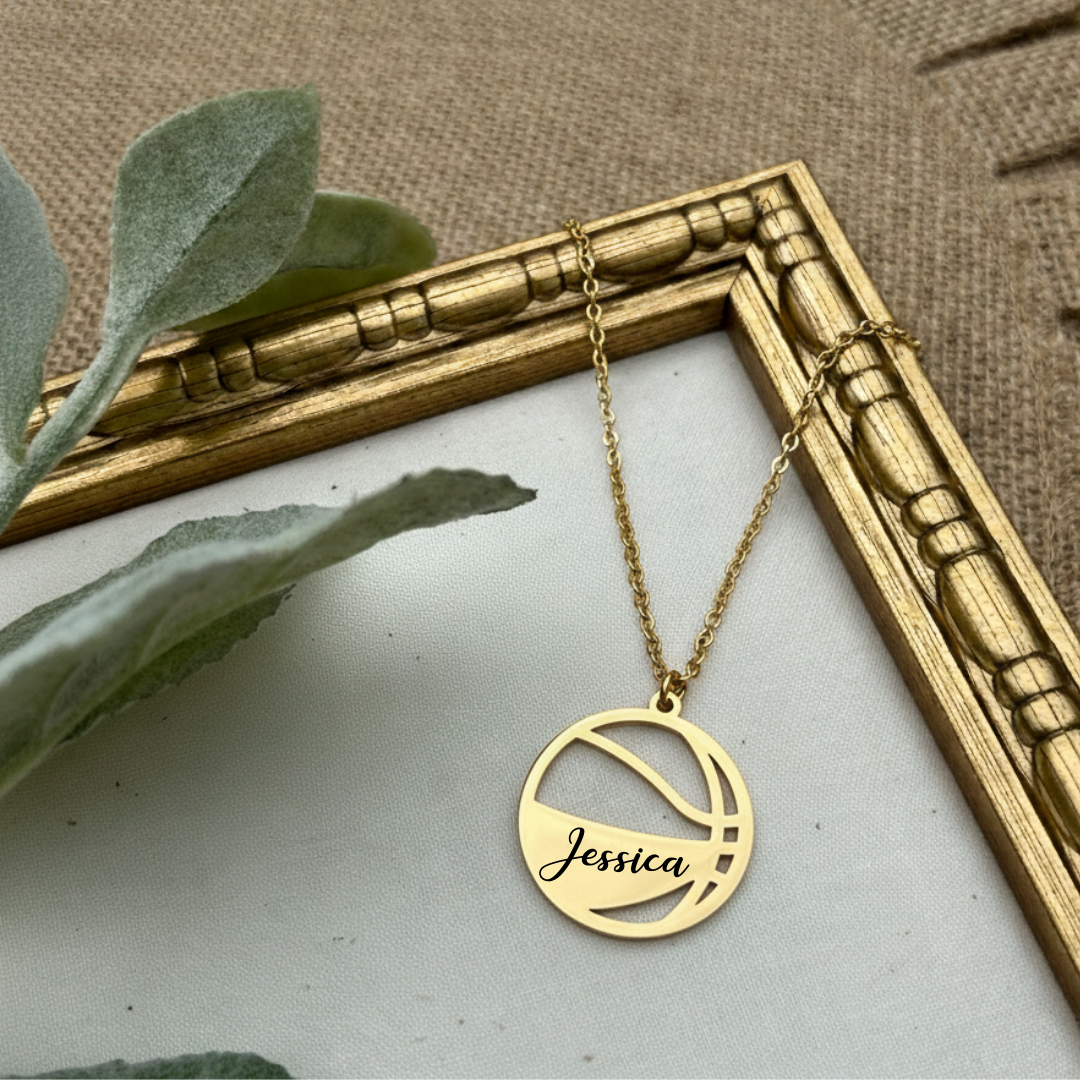 Gold necklace with 'Jessica' on the basketball pendant on a textured surface with a gold frame and green leaves.