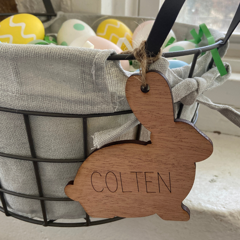 Wooden bunny tag with 'Colten' engraved, hanging from a wire basket with Easter eggs.