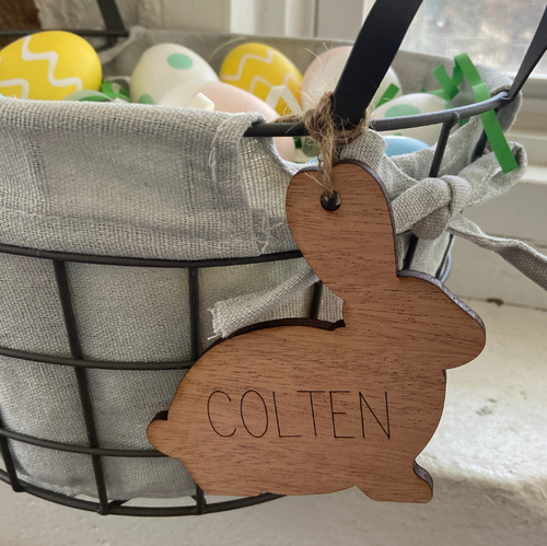 Wooden bunny tag with 'Colten' engraved, hanging from a wire basket with Easter eggs.