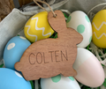 Wooden bunny-shaped ornament with 'COLTEN' engraved, surrounded by colorful Easter eggs.