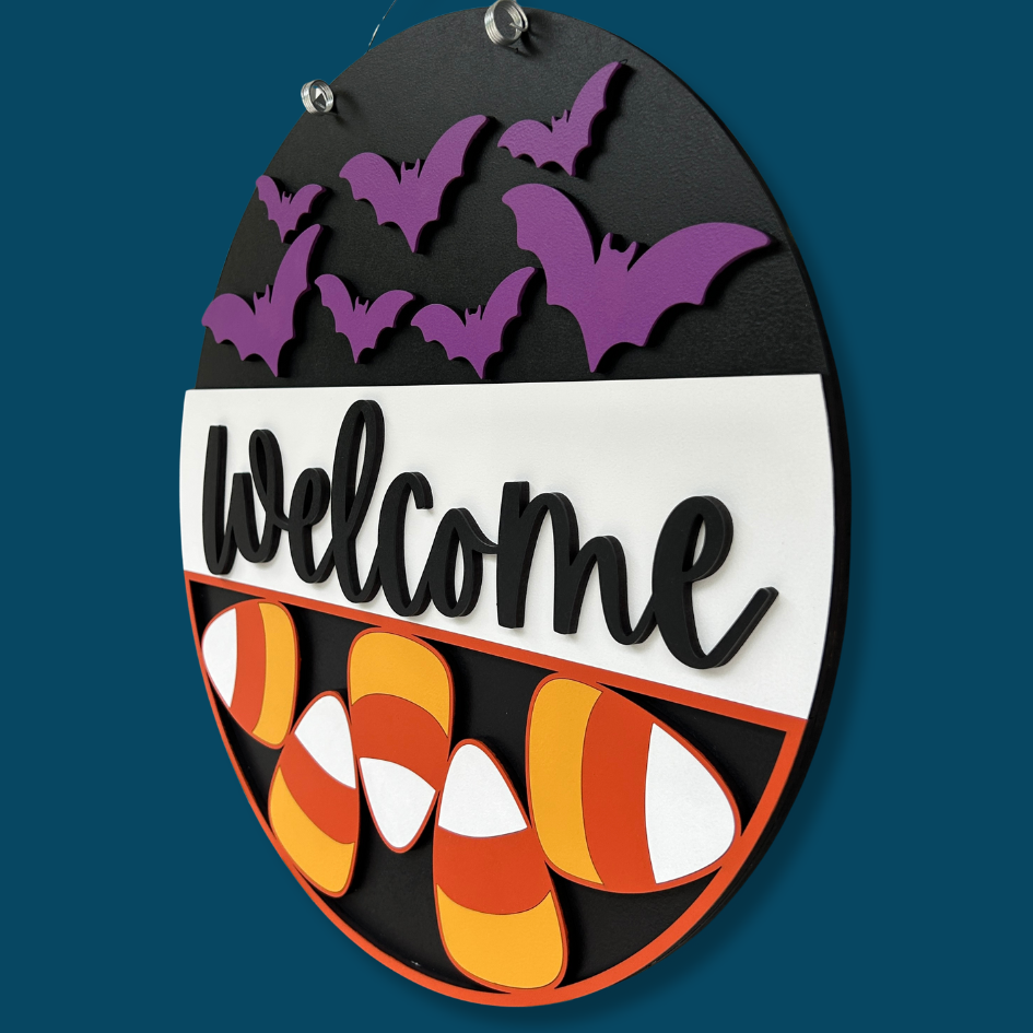 Halloween-themed decorative sign with purple bats, candy corn, and 'Welcome' text on a black background.