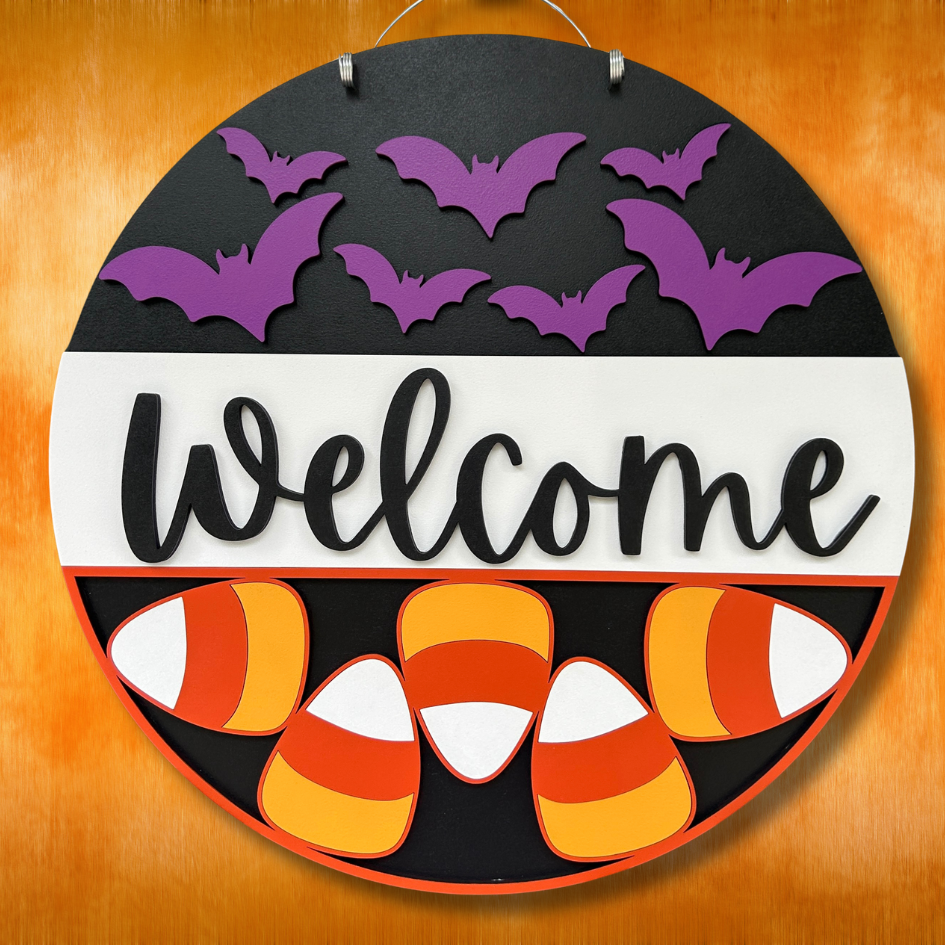 Halloween-themed decorative sign with purple bats, candy corn, and 'Welcome' text on a black background.