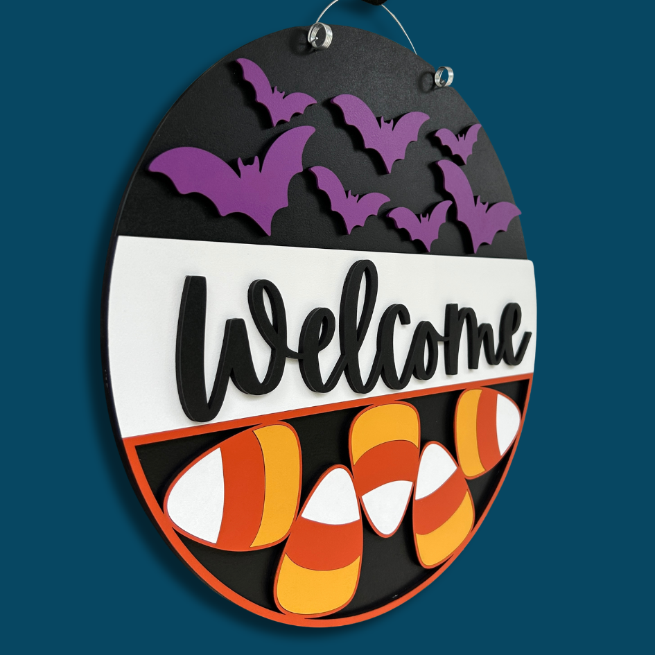 Halloween-themed decorative sign with purple bats, candy corn, and 'Welcome' text on a dark blue background.