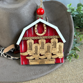Wooden Christmas ornament shaped like a barn with engraved names and 'Merry Christmas 2025' text, hanging from a hat.