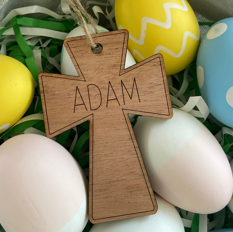 Wooden cross-shaped tag with 'ADAM' engraved, surrounded by colorful Easter eggs.