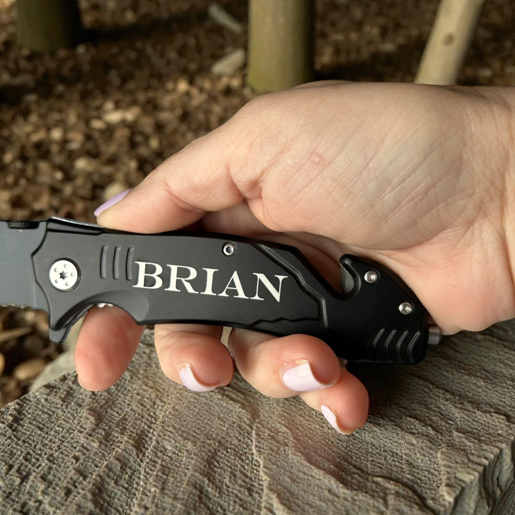 Hand holding a black folding knife with 'BRIAN' engraved on it, outdoors.