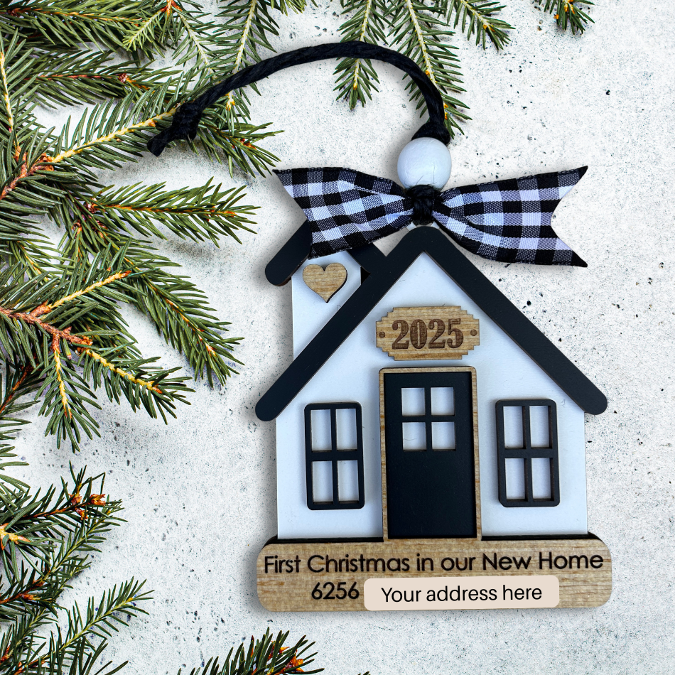 House-shaped Christmas ornament with 'First Christmas in our New Home' text and decorative bow, surrounded by pine branches.