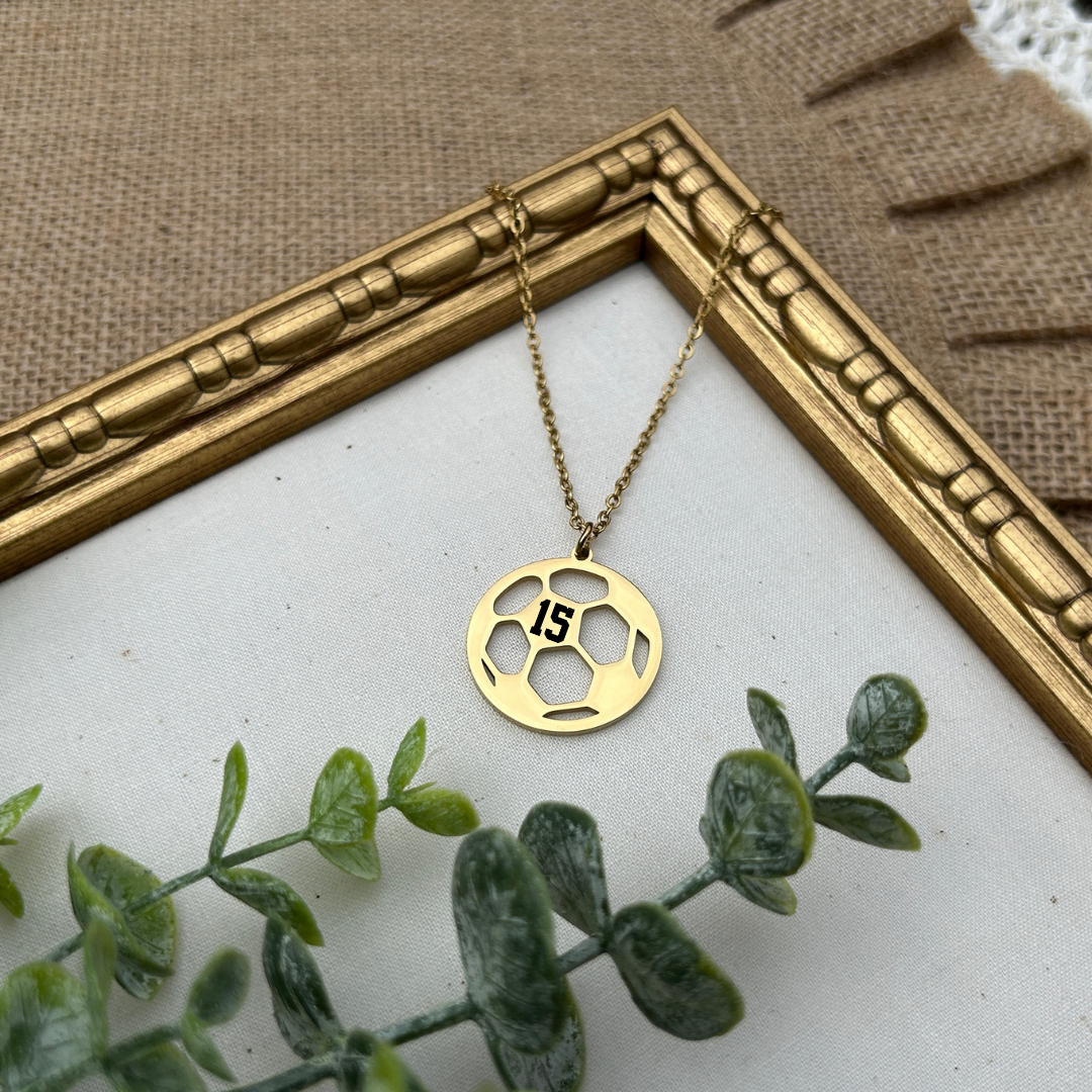 Gold necklace with a soccer ball pendant on a white surface with green leaves and a gold frame.