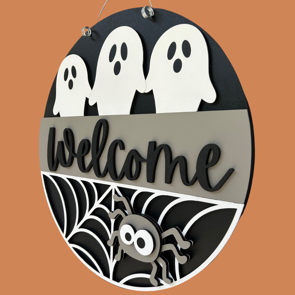 Halloween-themed decorative sign with ghosts, spider, and 'Welcome' text on a brown background.
