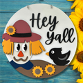 Decorative sign with a scarecrow and bird, featuring the text 'Hey Yall', on a blue wooden background.