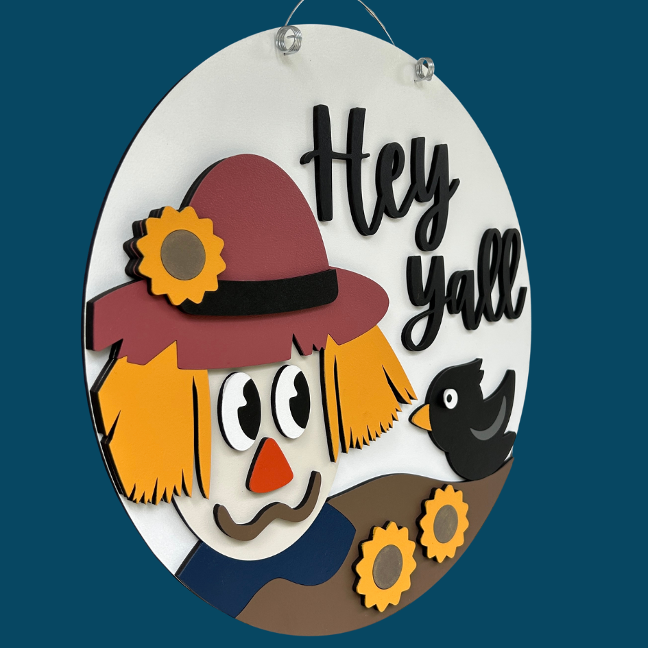 Decorative sign with a scarecrow face and 'Hey Yall' text on a blue background