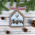 Decorative ornament with 'our first Christmas in our new home' text on a wooden background with pine branches and cones.