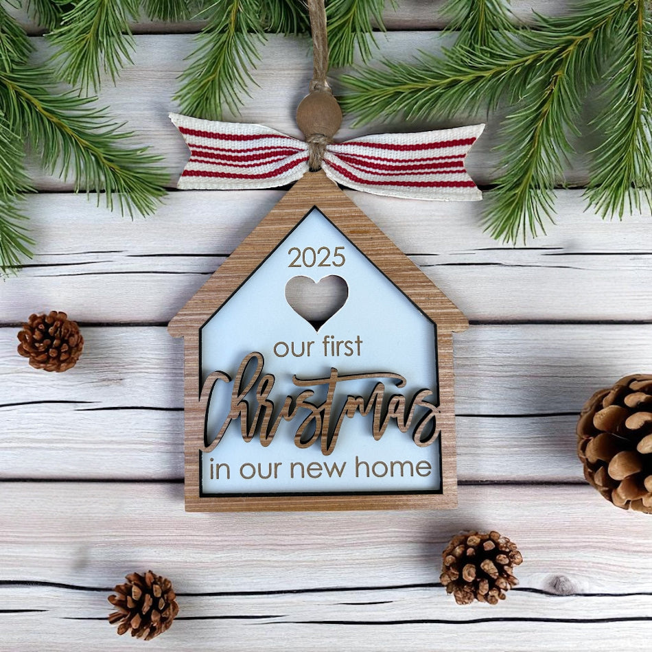 Decorative ornament with 'our first Christmas in our new home' text on a wooden background with pine branches and cones.