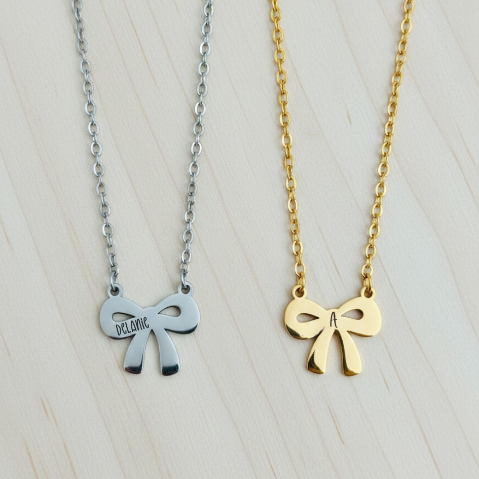 Two bow-shaped necklaces, one silver and one gold, on a light wooden surface.