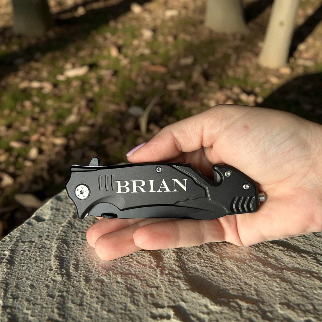 Hand holding a black folding knife with 'BRIAN' engraved on it, outdoors.