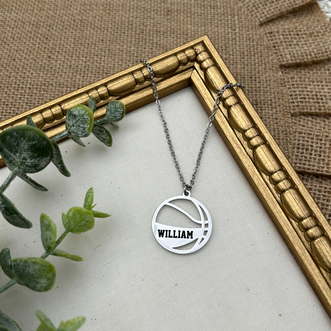 Silver necklace with a round basketball pendant displaying the name 'WILLIAM' on a textured surface with a gold frame and green leaves.