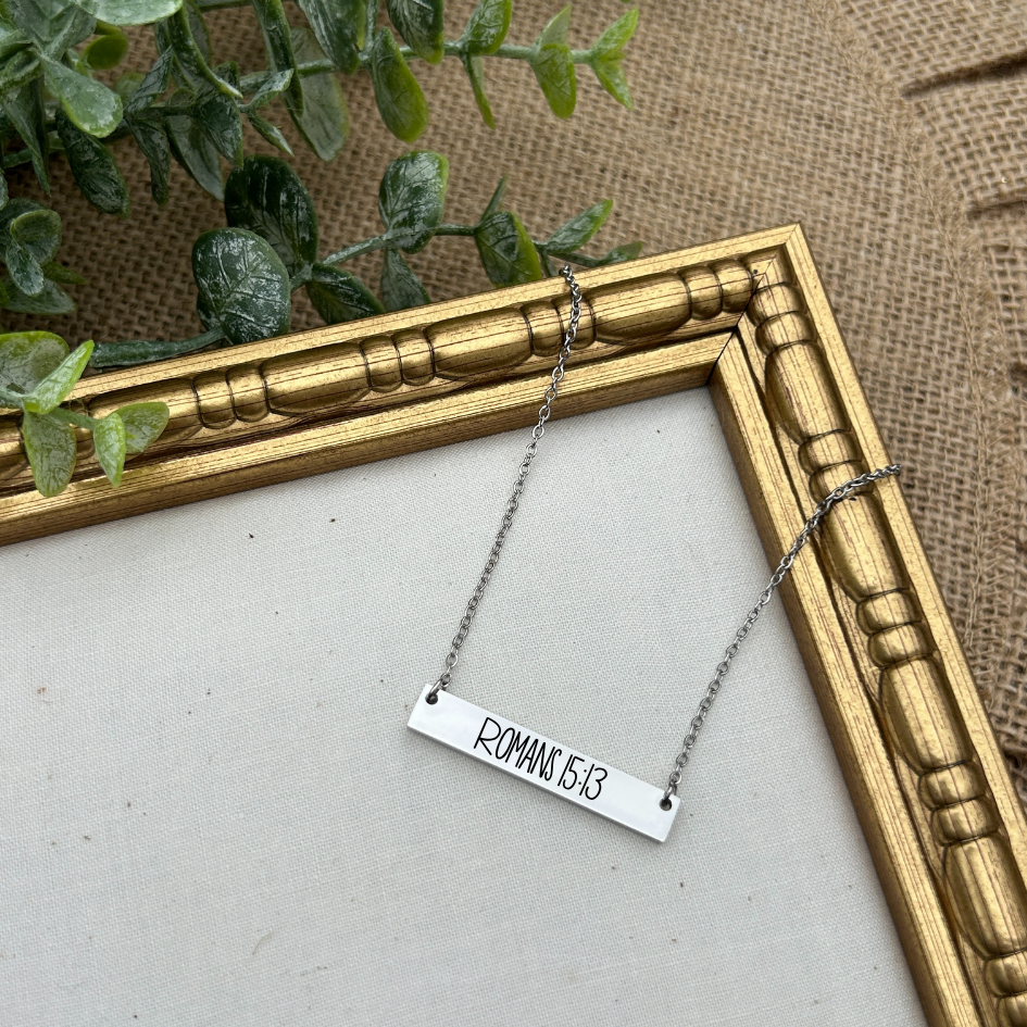 Necklace with 'Romans 15:13' pendant on a white surface with a gold frame and greenery in the background