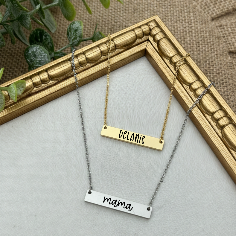 Two nameplate necklaces, one gold and one silver, on a light gray surface with a decorative gold frame in the background.