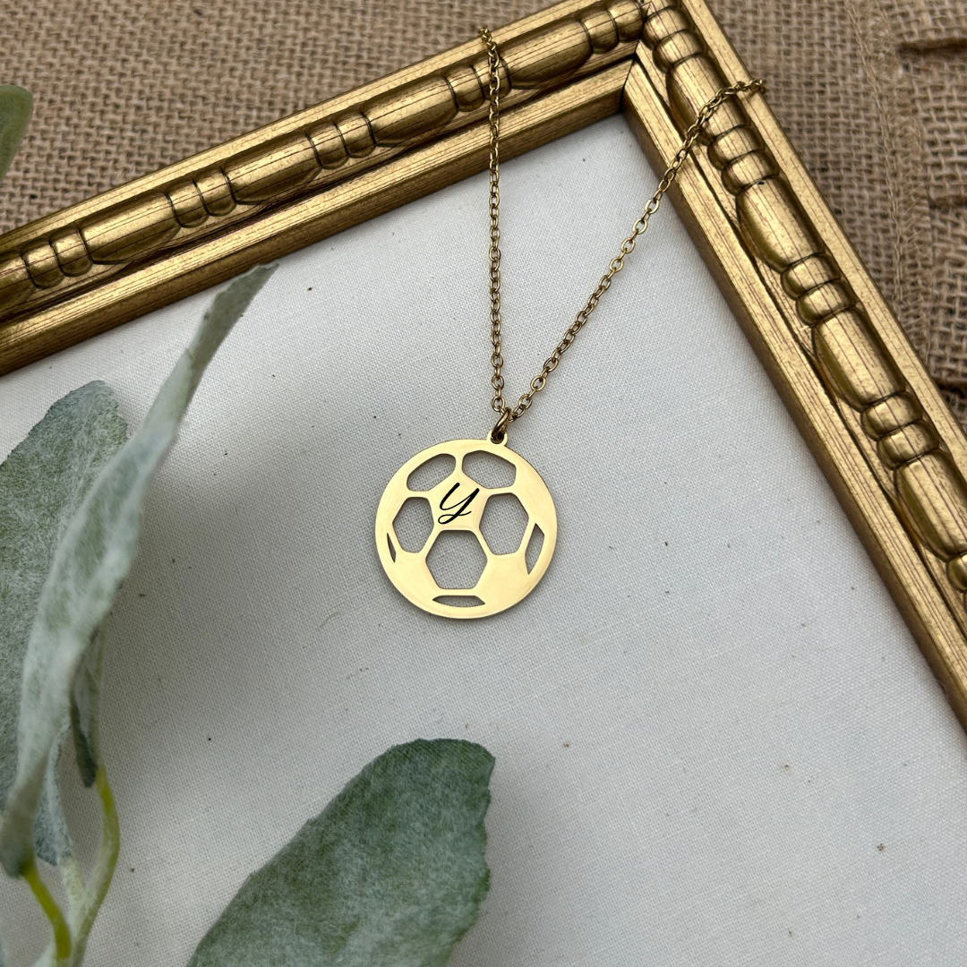 Gold necklace with a circular soccer pendant on a white surface with green leaves and a gold frame.