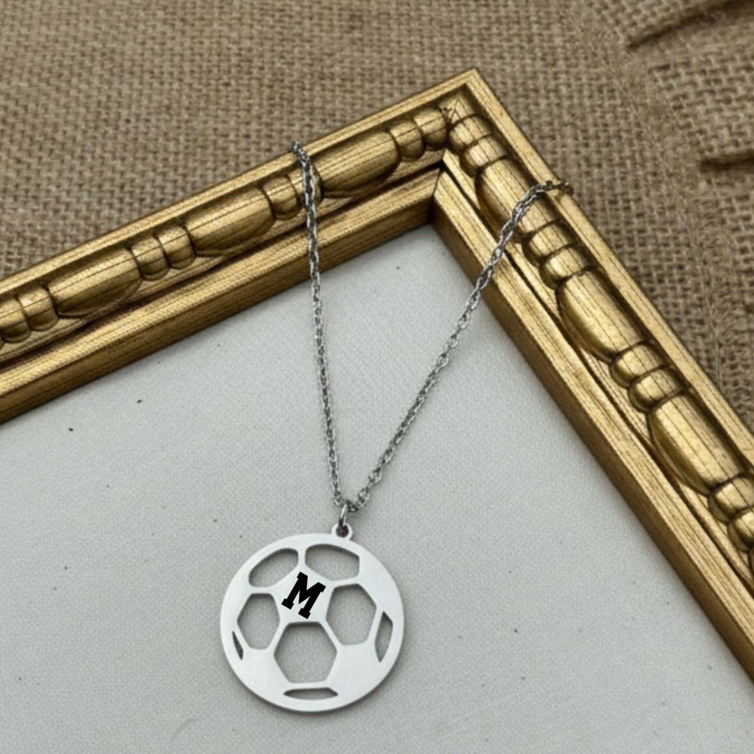 Silver necklace with a soccer pendant featuring a letter 'M' on a textured surface.