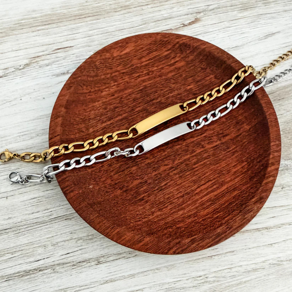 Two chain bracelets, one gold and one silver, on a wooden coaster.