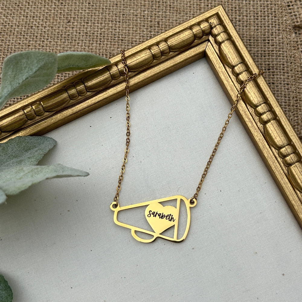 Gold necklace with a pendant shaped like a cheer megaphone on a textured surface with green leaves.