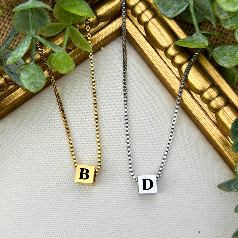 Two necklaces with 'B' and 'D' pendants on a textured surface with a gold frame and green leaves.
