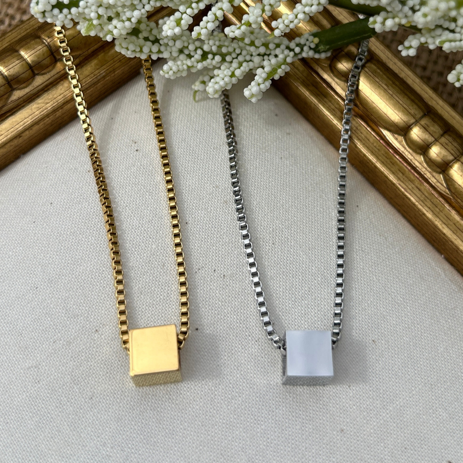 Two necklaces with square pendants on a textured surface with a gold frame and white flowers in the background.