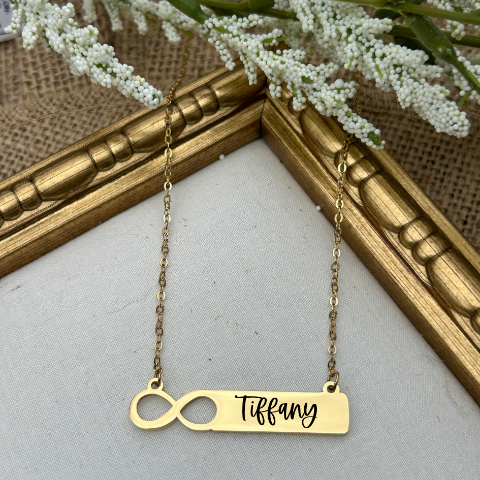 Gold necklace with 'Tiffany' pendant on a white surface with a gold frame and white flowers in the background