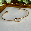Gold bracelet with knot design on a light gray surface with greenery in the background