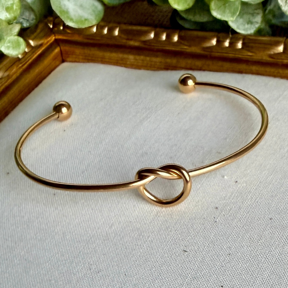 Gold bracelet with knot design on a light gray surface with greenery in the background