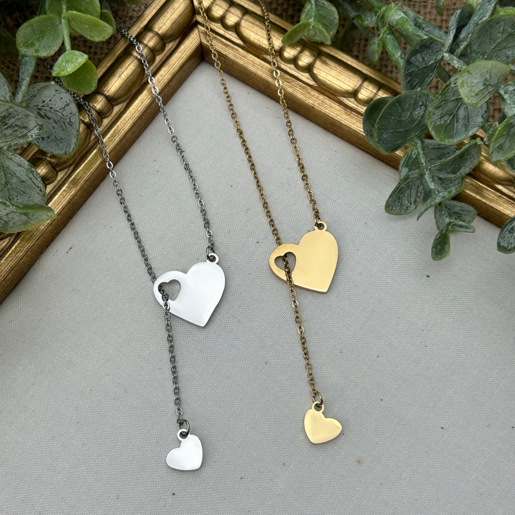 Two heart-shaped necklaces, one silver and one gold, on a textured surface with greenery in the background.