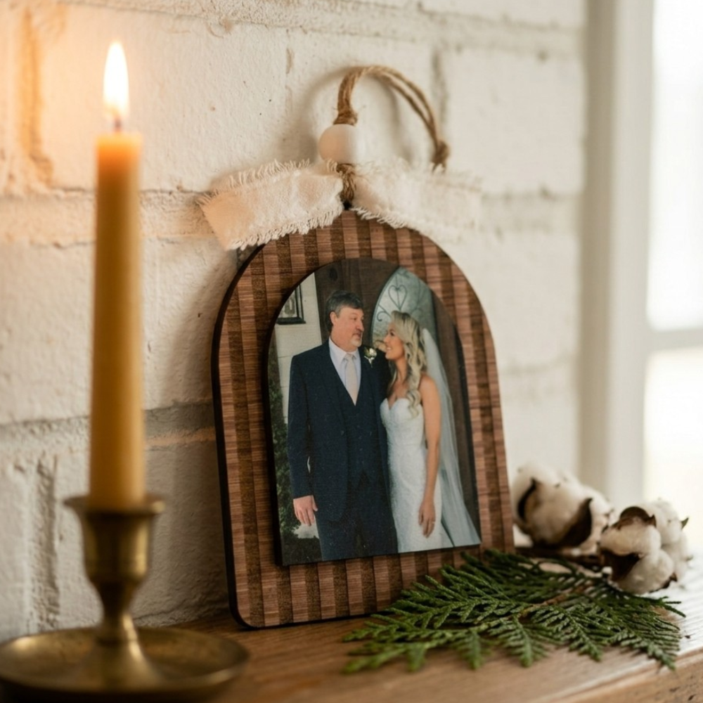 Decorative wooden frame with a photo of a father and daughter, lit candle, and greenery on a rustic surface.