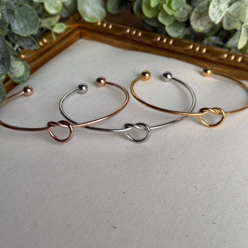 Three gold, silver, and rose gold bangles on a textured surface with greenery in the background.