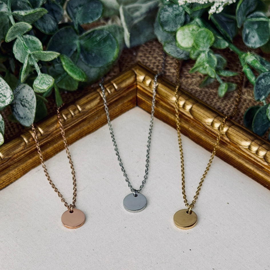 Three necklaces with round pendants on a textured surface with plants in the background