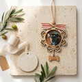 Wooden ornament with a dog's face, surrounded by greenery and ribbons on a textured surface.