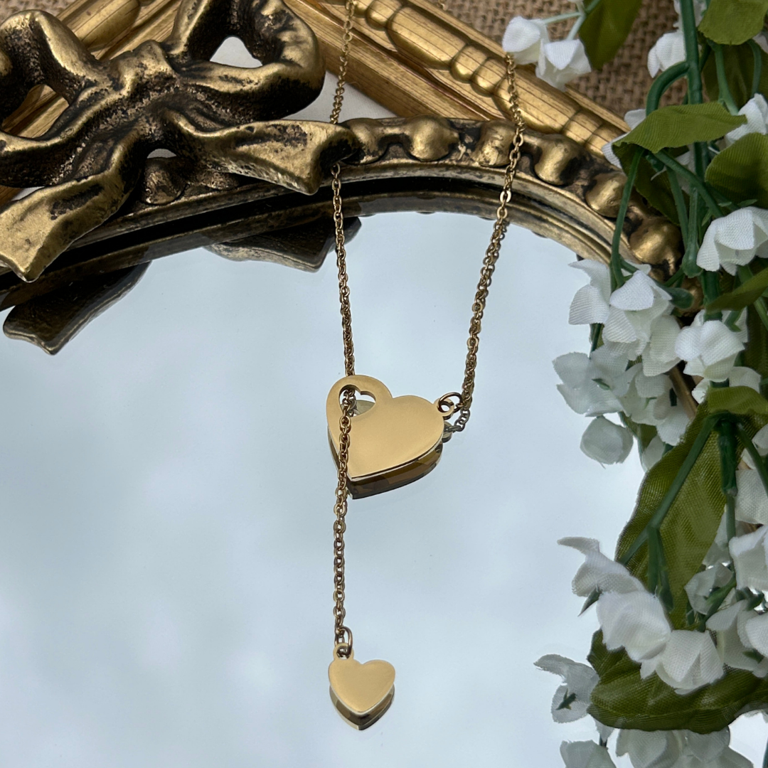 Gold heart-shaped pendant on a chain with a decorative gold frame and white flowers in the background.