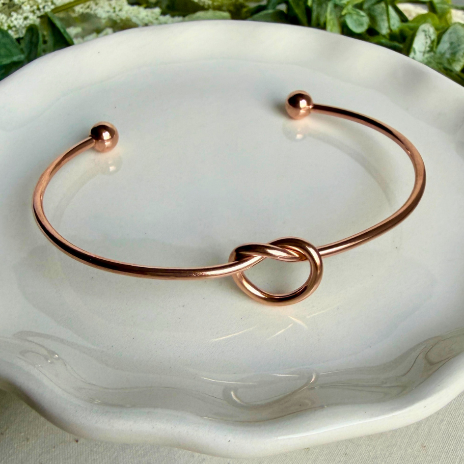 Rose gold bracelet with knot design on a white dish with greenery in the background