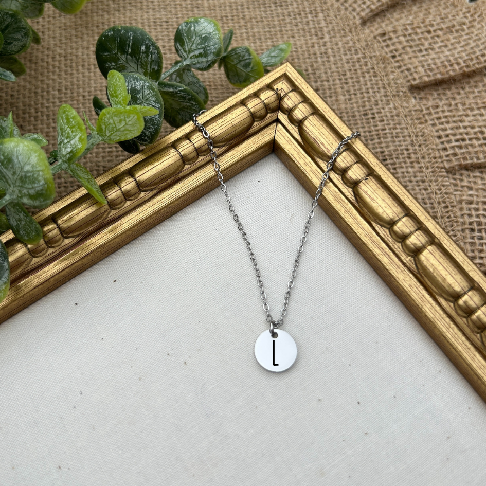Necklace with a round pendant featuring the letter 'L' on a textured surface with greenery.