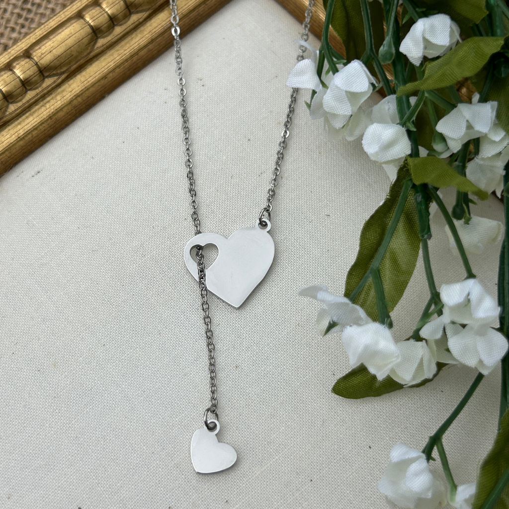 Silver heart-shaped necklace on a neutral background with white flowers and green leaves.