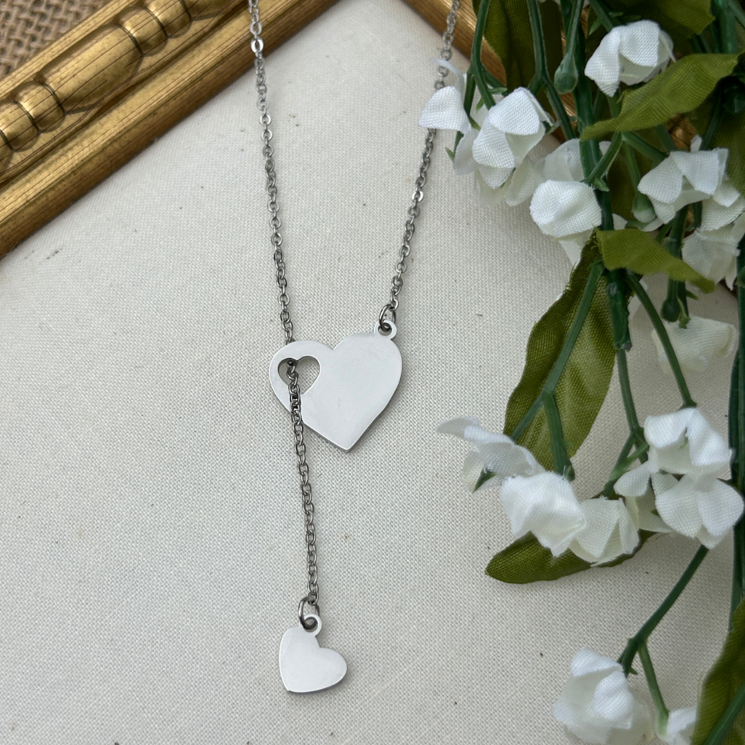 Silver heart-shaped necklace on a neutral background with white flowers and green leaves.