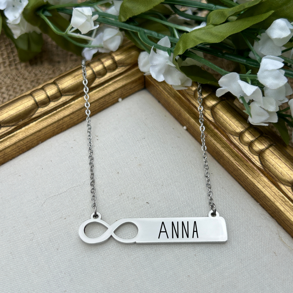 Silver necklace with 'ANNA' pendant on a neutral background with flowers and a gold frame.