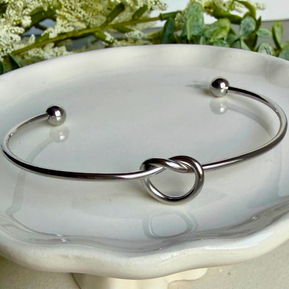 Silver bracelet with a knot design on a white dish with greenery in the background