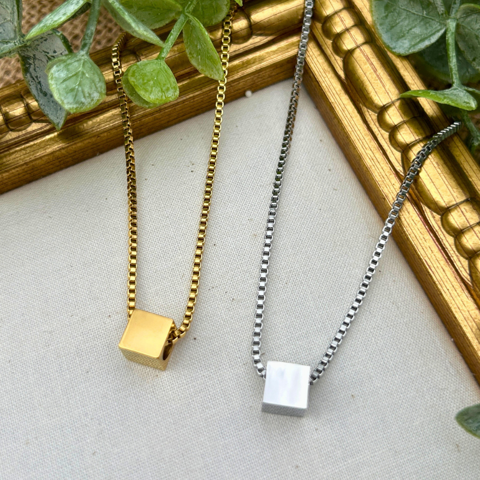 Two necklaces with square pendants on gold and silver chains, placed against a gold frame with greenery.