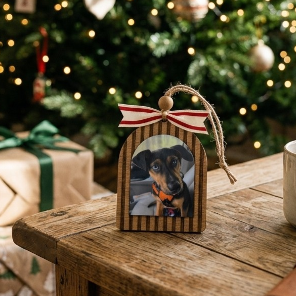 Wooden ornament with a photo of a dog on a wooden table with a Christmas tree in the background.